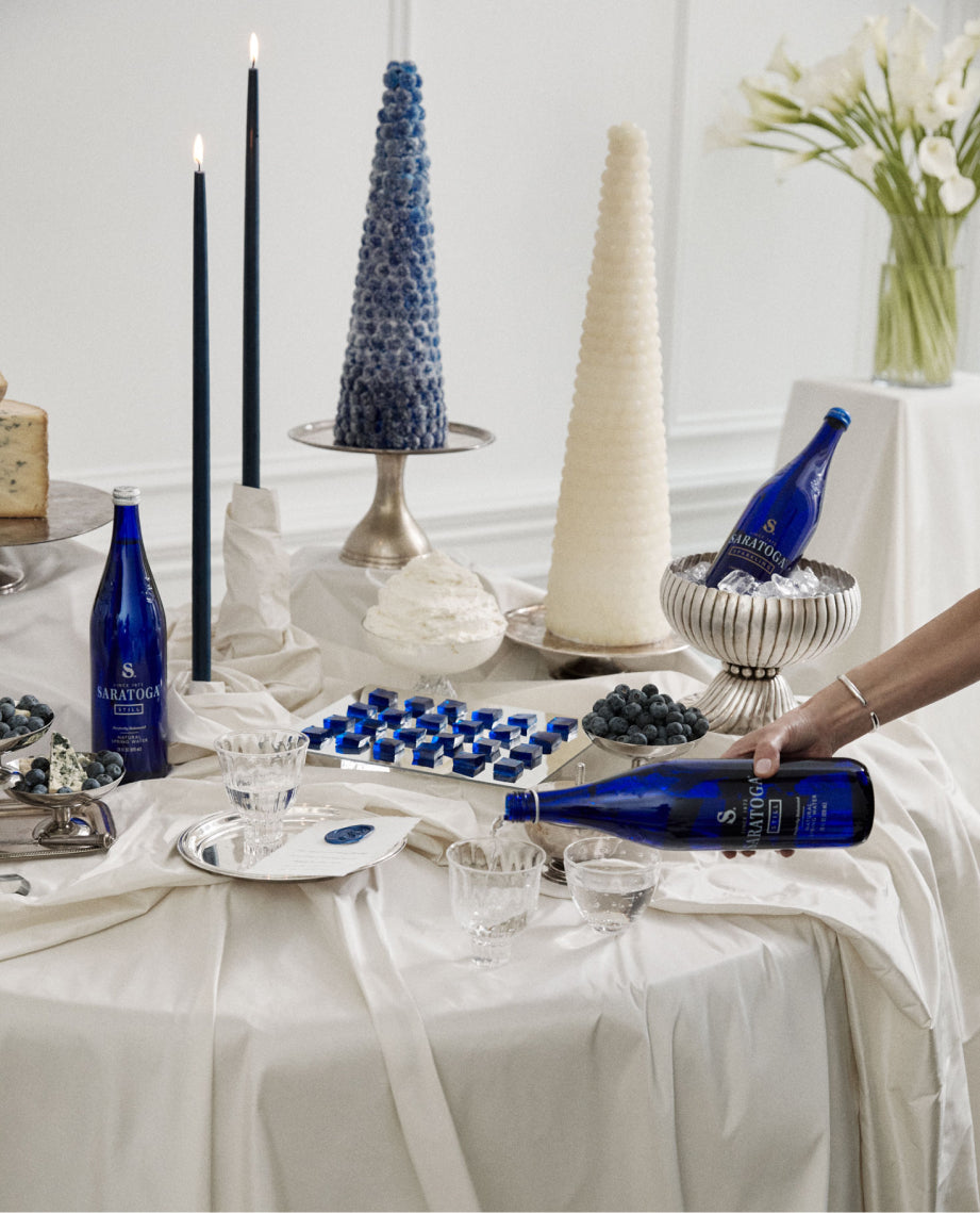 woman pouring saratoga spring water into a crystal goblet on a table decorated with blue candles and blue desserts