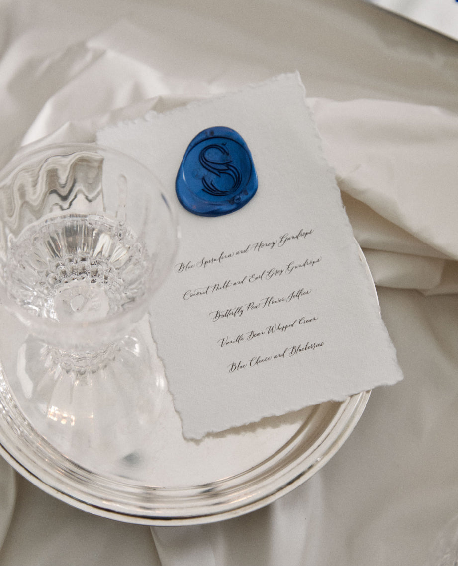 elegant cursive handwritten menu with blue wax stamp of saratoga water brand logo laid on a silver platter next to a crystal drinking glass